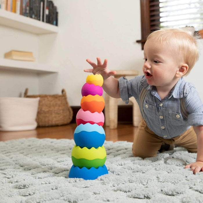 Toddler reaching for a stack of six colorful, weighted, sphere-like toys with wavy edges on a plush gray rug.