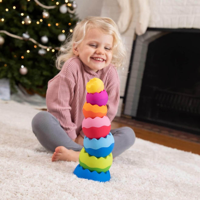 Child smiles at a stack of seven vibrant, textured spheres in rainbow colors on a rug near a Christmas tree.