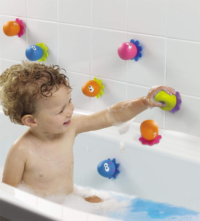A toddler in a bubble bath sticks colorful, numbered octopus suction toys with smiling faces onto white wall tiles.
