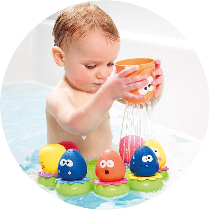 A baby pouring water from an orange octopus cup onto small, colorful octopus toys on a floating green base in a bath.