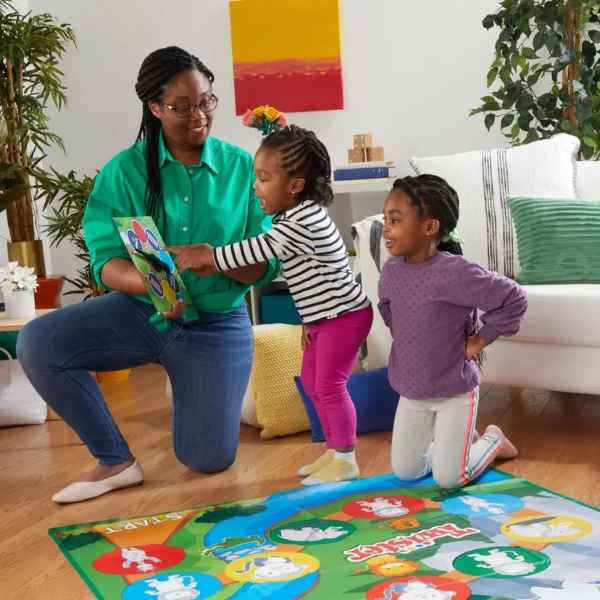 Adult and children play Twister Junior on a colorful jungle-themed floor mat with a handheld game spinner.
