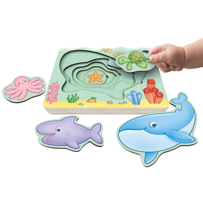Child fits a green turtle piece into a layered wooden ocean puzzle with a pink octopus, purple shark, and blue whale.