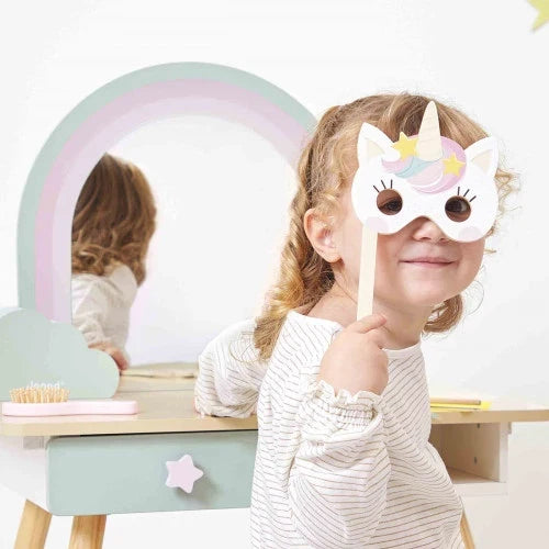 Girl holding a unicorn mask at a pastel rainbow wooden vanity with a star-knobbed drawer and matching hairbrush.