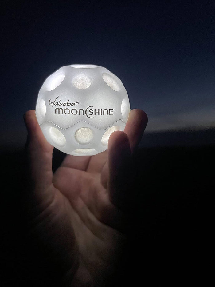 Hand holding a glowing white ball with crater dimples and black Moonshine text against a dark background.