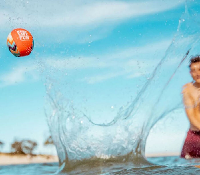 Orange and blue geometric patterned ball leaping out of a large water splash against a bright blue sky.