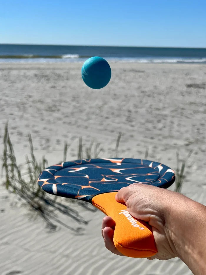 Hand holds a navy and orange patterned fabric paddle with a blue ball mid-air on a sunny sandy beach.