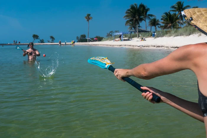 Two people playing catch with blue and yellow lacrosse sticks and an orange ball in clear ocean water at a sunny beach.