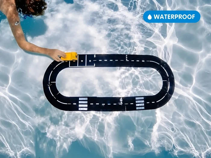 A child underwater pushing a yellow toy car on a black oval road track at the bottom of a clear blue pool.