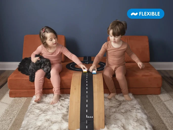 Two children racing toy cars on flexible black road tracks over an orange play sofa and a wooden balance board ramp.
