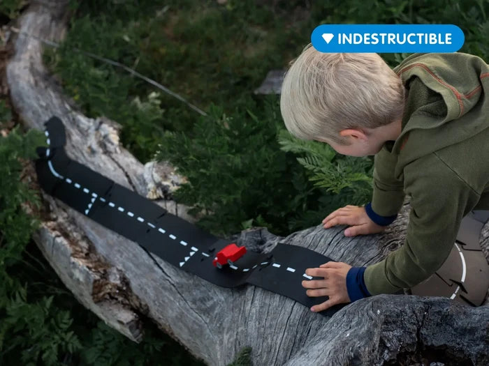 Child playing with flexible black toy road pieces on a fallen tree log with a small red car.