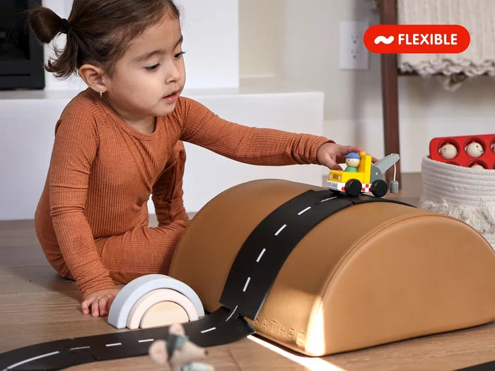 Flexible black rubber road track with white dashed lines draped over a tan foam arch being used by a child.