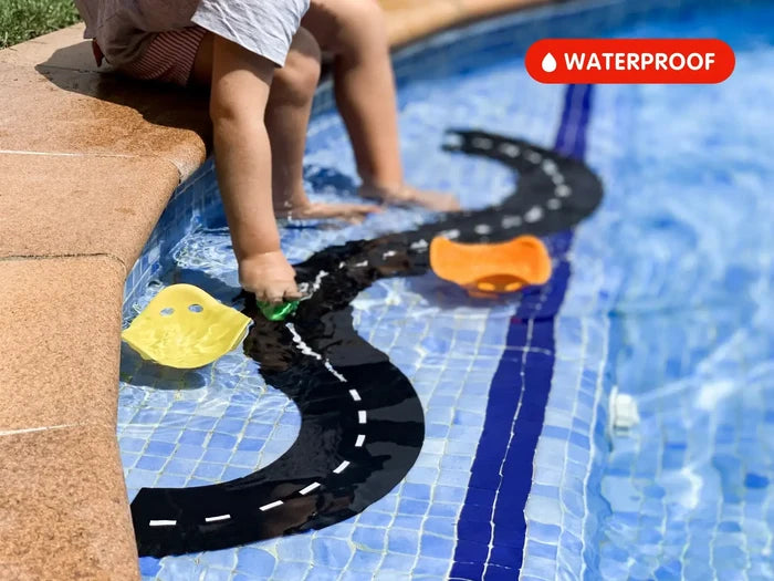 Black flexible toy road tracks with white markings floating in a blue pool as a child plays. Red waterproof label shown.