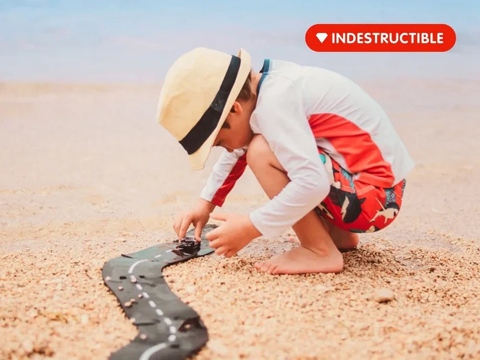 Child playing with a toy car on a flexible black road with white lines, laid out on a pebbly beach.