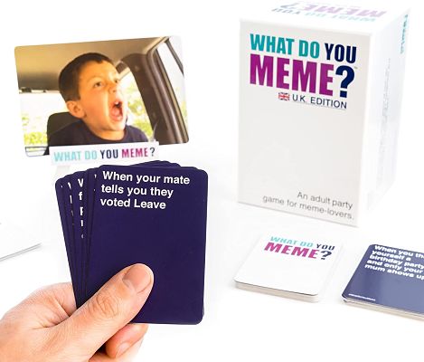 What Do You Meme? UK Edition game box with photo cards and a hand holding purple caption cards on a white background.