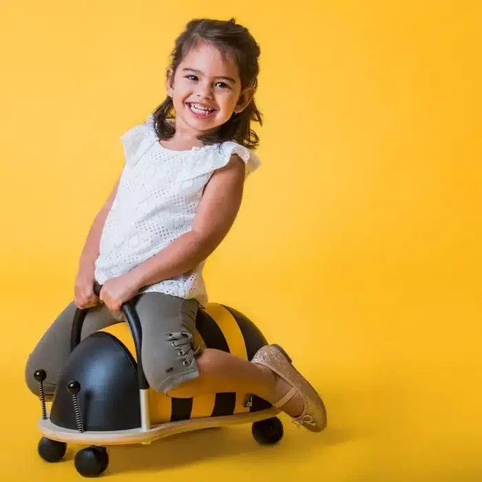 Smiling child on a yellow and black striped bee ride-on toy with a handle and caster wheels on a yellow background.