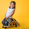 Smiling child on a yellow and black striped bee ride-on toy with a handle and caster wheels on a yellow background.