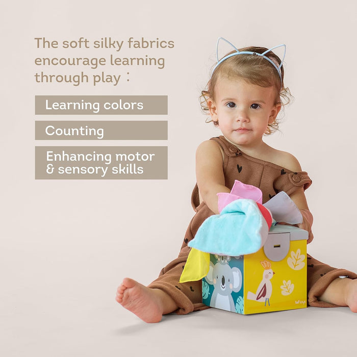 Toddler girl pulls colorful fabrics from an illustrated yellow box; text highlights motor and sensory skills.