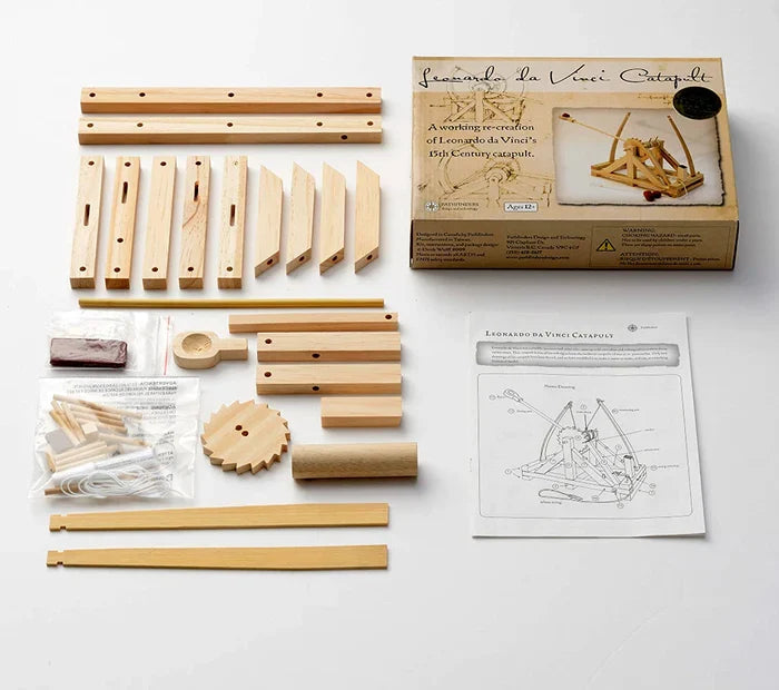 Unassembled light wood catapult kit with beams, gears, instructions, and a retail box on a white background.