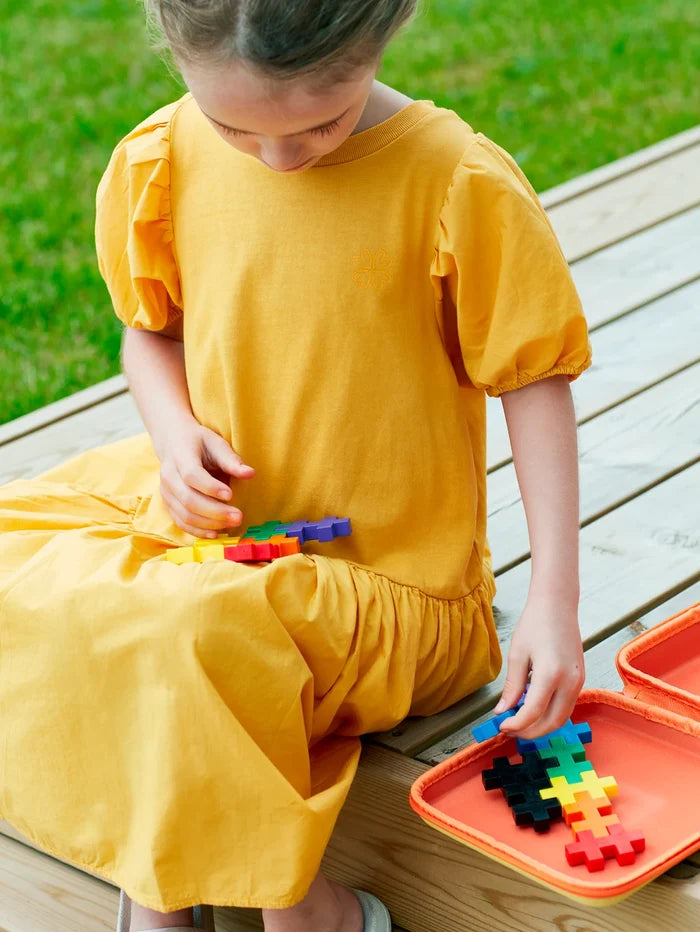 Child playing with colorful plus-shaped building blocks from an orange zippered travel case on a wooden deck.