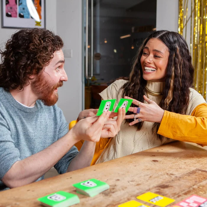 Man holding green cards with the letters B and A while laughing with a woman during a card game at a wooden table.