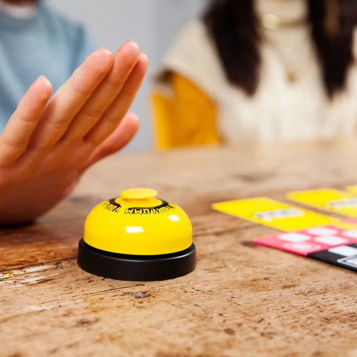 Hand poised to press a yellow call bell next to colorful quiz cards on a rustic wooden table.