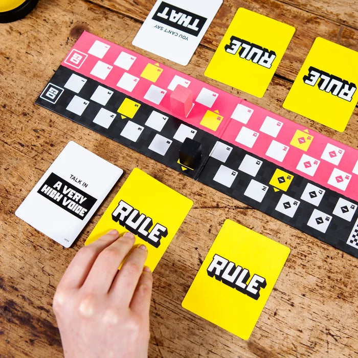 Game boards with pink and black paths, yellow RULE cards, and a hand picking a card on a wooden table.