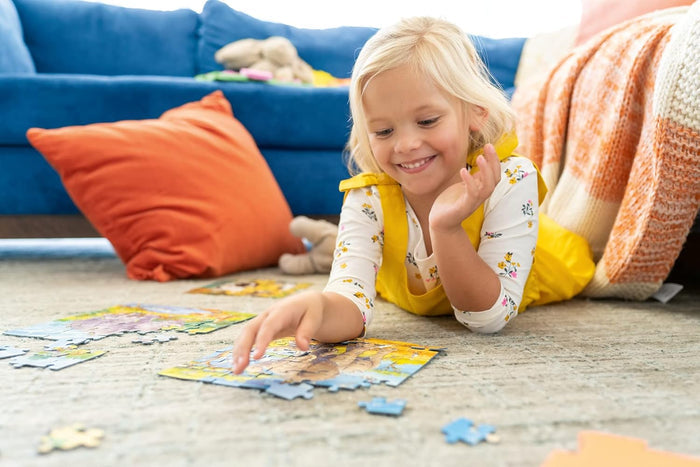 A young girl in a yellow dress smiles while assembling colorful animal-themed jigsaw puzzles on a light-colored rug.
